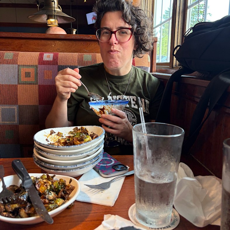 A woman holding a sundae glass enjoying a brownie sundae with five empty plates piled in front of her and some portions of brussel sprouts